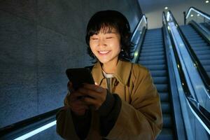 sorridente coreano menina indo baixa escada rolante dentro escuro, segurando Móvel telefone, usando Smartphone aplicativo, ouvindo música, pendulares dentro cidade foto