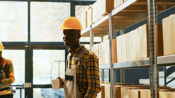 africano americano armazém trabalhador posando perto prateleiras preenchidas com produtos dentro caixas. jovem homem com macacão e capacete trabalhando dentro armazenamento quarto depósito com varejo loja mercadoria. foto