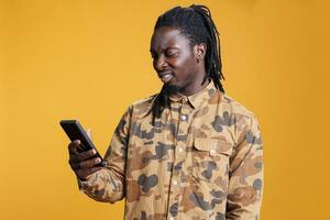 africano americano homem lendo trabalho inscrição em Smartphone, rolagem através social meios de comunicação dentro estúdio sobre amarelo fundo. alegre jovem adulto procurando em formação em Móvel telefone foto