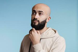 árabe homem olhando longe com pensativo facial expressão e fricção queixo pensativo. jovem Careca barbudo pessoa pensando e fazer decisão enquanto posando com intrigado gesto foto