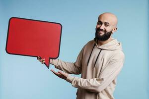 alegre árabe homem promovendo Novo produtos com quadrinho balão vermelho bandeira retrato. sorridente pessoa segurando esvaziar diálogo bolha e olhando às Câmera com feliz facial expressão foto