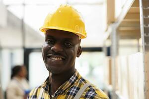 africano americano armazém pacote manipulador dentro amarelo capacete retrato. industrial armazém jovem carregador vestindo protetora vestuário de trabalho sorridente e olhando às Câmera fechar acima foto