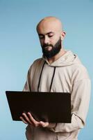 concentrado árabe homem lendo o negócio relatório em computador portátil e digitando conectados mensagem. focado jovem pessoa segurando portátil computador, usando Programas apps e navegando Internet foto