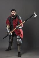 cavaleiro medieval em fundo cinza. retrato de guerreiro brutal com rosto sujo com cota de malha, roupas vermelhas e pretas e machado de batalha foto