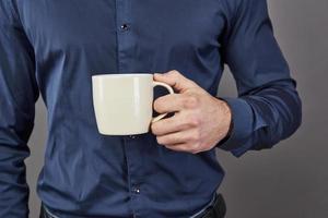 bonito homem barbudo com cabelo elegante, barba e bigode na cara séria na camisa segurando um copo branco ou caneca, bebendo chá ou café no estúdio em fundo cinza foto