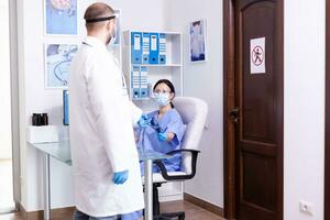 médico vestindo protetora casaco e face mascarar contra coronavírus enquanto dando enfermeira paciente radiografia dentro hospital esperando sala. assistente trabalhando em recepção computador. foto