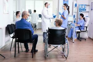 Desativado Senior mulher vestindo face mascarar contra coronavírus dentro hospital esperando área e tendo uma conversação com velho cara. assistente trabalhando em recepção computador. foto