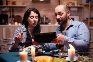 alegre jovem casal jantar juntos usando tábua durante romântico jantar. adultos sentado às a mesa dentro a cozinha navegando, procurando, , Internet, a comemorar, aniversário, feliz., festivo, apreciar. foto