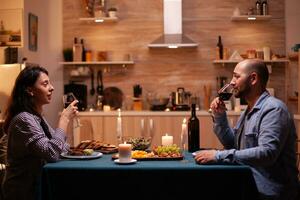 casal desfrutando uma vidro do vinho. relaxar feliz pessoas tinindo, sentado às mesa dentro cozinha, desfrutando a refeição, a comemorar aniversário dentro a jantar sala. foto