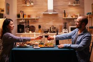 positivo marido e esposa segurando vinho óculos dentro cozinha dentro a tarde. relaxar feliz pessoas tinindo, sentado às mesa dentro cozinha, desfrutando a refeição, a comemorar aniversário dentro a jantar sala. foto