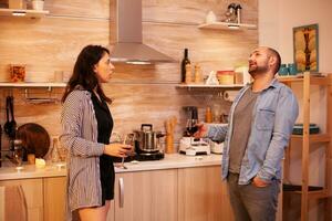 esposa dizendo uma história para dela marido enquanto segurando uma vidro do vinho dentro cozinha. adulto casal às lar, bebendo vermelho vinho, conversando, sorridente, desfrutando a refeição dentro jantar sala. foto