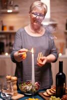 Senior mulher com óculos iluminação vela em cozinha mesa para romântico jantar. idosos mulher esperando dela marido para uma romântico jantar. maduro esposa preparando festivo refeição para aniversário celebração. foto