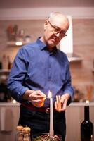 feliz Senior marido usando fósforos luz velas para romântico aniversário com esposa. idosos velho marido preparando festivo refeição com saudável Comida para aniversário celebração, sentado perto a mesa. foto