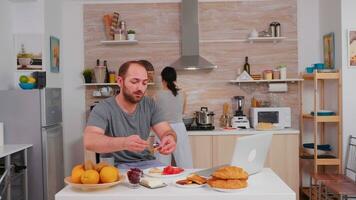 empreendedor trabalhando em computador portátil dentro cozinha e bebendo café enquanto esposa é cozinhando café da manhã. trabalhador autonomo trabalhos conectados através da Internet usando moderno digital tecnologia foto