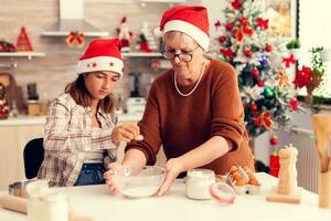 amoroso avó em Natal dia fazer biscoitos com sobrinha. feliz alegre alegre Adolescência menina ajudando Senior mulher preparando doce biscoitos para comemoro inverno feriados vestindo santa chapéu. foto