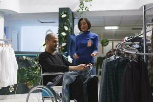 inclusivo roupas loja ásia mulher assistente fornecendo orientação para cliente dentro cadeira de rodas. árabe homem dentro cadeira de rodas comprando vestuário e obtendo moda adendo a partir de boutique vendedor foto