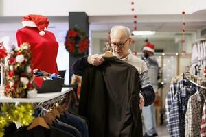 Senior cliente navegando jaquetas dentro festivo roupas loja durante inverno Natal feriado temporada. pessoa olhando às sazonal roupas em desconto, fazendo varejo terapia para natal festividade apresenta. foto