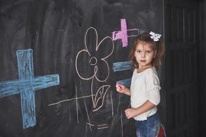 garotinha encaracolada desenhando com giz de cera na parede. obras de criança. aluno fofo escrevendo no quadro-negro foto