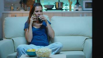 mulher dentro pijamas falando às telefone assistindo televisão e comendo lanches. feliz, relaxado, solitário senhora dentro pijamas desfrutando a tarde sentado em confortável sofá dentro frente do televisão Falando em Smartphone foto