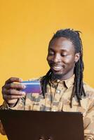 africano americano homem usando crédito cartão para fazer compras conectados usando Smartphone moda roupas aplicativo, encomenda à moda roupas. jovem adulto pagando produtos em digital loja dentro estúdio sobre amarelo fundo foto