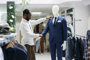 boutique assistente verificação manequim vestido dentro formal roupa masculina Mais recentes coleção. africano americano homem examinando masculino luxo terno em modelo, enquanto trabalhando dentro compras Shopping foto