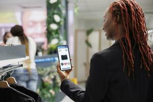 jovem cliente comprando roupas em conectados boutique aplicativo, olhando às na moda roupas linha em Móvel telefone local na rede Internet. fêmea comprador escolhendo moda Itens em e comércio inscrição dentro fazer compras. foto