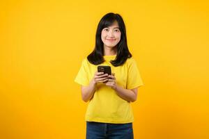 experiência a felicidade e conexão do tecnologia com alegre retrato. jovem ásia mulher vestindo amarelo camiseta e jeans jeans sorrisos usando Smartphone. conceito do tecnologia e Smartphone aplicativo. foto