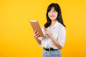 jovem ásia mulher vestindo branco camisa sorrisos brilhantemente enquanto segurando digital tábua contra amarelo fundo, representa uma confiante companhia trabalhador abraçando moderno tecnologia conceitos. foto