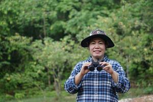 ásia mulher explorador desgasta chapéu, azul xadrez camisa , detém binóculos para explorar, pesquisa botânico plantas e criaturas animais selvagens. conceito, natureza exploração. ecologia e ambiente. foto