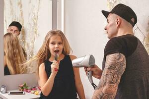 fofa pequeno filha e dela tatuado Papai estão jogando juntos perto uma espelho. Papai é fazendo dele da filha cabelo. família feriado e união. foto