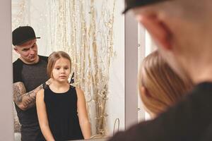 fofa pequeno filha e dela tatuado Papai estão jogando juntos perto uma espelho. Papai é fazendo dele da filha cabelo. família feriado e união. foto