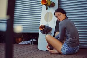 morena menina dentro cinzento gola alta e jeans calção é posando com dela prancha de wakeboard sentado em chão às uma costeiro zona. esporte e lazer. fechar-se. foto