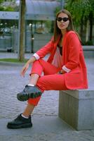 Loiras menina dentro uma vermelho tipo senhora terninho e Preto principal, assistir, pingente, oculos de sol é posando sentado em uma pedra banco. moda conceito. comprimento total. foto