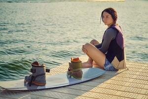 morena menina dentro uma cinzento gola alta, especial Esportes colete e azul jeans calção é posando com dela prancha de wakeboard em uma cais do a costeiro zona. fechar-se. foto