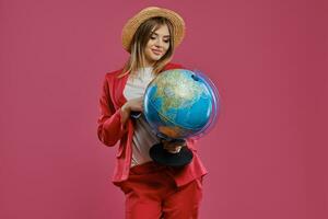 Loiras fêmea dentro Palha chapéu, branco blusa e vermelho terninho. ela é sorridente, segurando uma globo, Passaporte e bilhete, posando em Rosa fundo. fechar-se foto