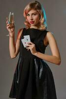 Loiras menina dentro Preto vestir segurando dois jogando cartões e vidro do champanhe, posando contra cinzento fundo. jogatina, pôquer, casino. fechar-se. foto