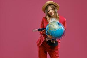 Loiras senhora dentro Palha chapéu, branco blusa e vermelho terninho. ela é sorridente, segurando globo, Passaporte e bilhete, posando contra Rosa fundo. fechar-se foto