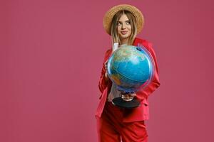 Loiras modelo dentro Palha chapéu, branco blusa e vermelho terninho. ela é sorridente, segurando globo, Passaporte e bilhete, posando contra Rosa fundo. fechar-se foto