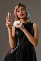 Loiras menina dentro Preto vestir segurando dois jogando cartões e vidro do champanhe, posando contra cinzento fundo. jogatina, pôquer, casino. fechar-se. foto