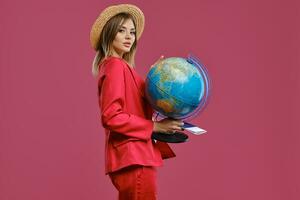 Loiras menina dentro Palha chapéu, branco blusa e vermelho terninho. ela é segurando uma globo, Passaporte e bilhete, posando lateralmente em Rosa fundo. fechar-se foto