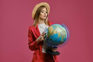 Loiras modelo dentro Palha chapéu, branco blusa e vermelho terninho. ela é segurando uma globo, Passaporte e bilhete enquanto posando contra Rosa fundo. fechar-se foto