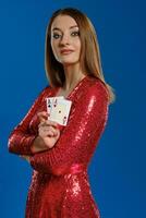 Loiras mulher com inventar, dentro vermelho lantejoulas vestir é mostrando dois ases, mãos guardada, posando em azul fundo. jogatina, pôquer, casino. fechar-se. foto