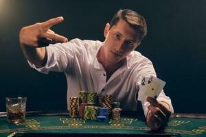 bonito emocional homem é jogando pôquer sentado às a mesa dentro cassino contra uma branco Holofote. foto