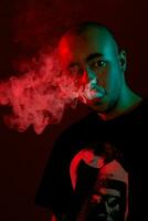 fechar-se estúdio tiro do uma jovem Careca cara Cigarro eletrônico, sopro Fora uma nuvem do fumaça em vermelho fundo. foto