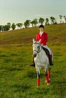 jovem mulher cavaleiro, vestindo vermelho redingote e branco calças, com dela cavalo dentro tarde pôr do sol claro. foto