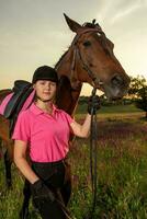 lindo sorridente menina jóquei ficar de pé Próximo para dela Castanho cavalo vestindo especial uniforme em uma céu e verde campo fundo em uma pôr do sol. foto