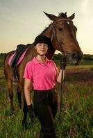 lindo sorridente menina jóquei ficar de pé Próximo para dela Castanho cavalo vestindo especial uniforme em uma céu e verde campo fundo em uma pôr do sol. foto