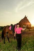 lindo sorridente menina jóquei ficar de pé Próximo para dela Castanho cavalo vestindo especial uniforme em uma céu e verde campo fundo em uma pôr do sol. foto