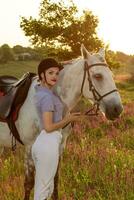 jóquei jovem menina acariciando e abraçando branco cavalo dentro tarde pôr do sol. Sol flare foto