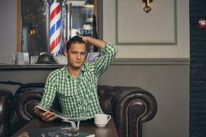 bonito jovem homem dentro uma barbearia é esperando para a mestre dentro a esperando quarto enquanto bebendo café foto